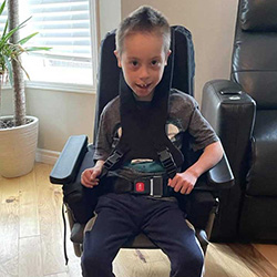 A young boy named Liam smiles while seated in a supportive pediatric wheelchair, designed for conditions like autism and cognitive disabilities.